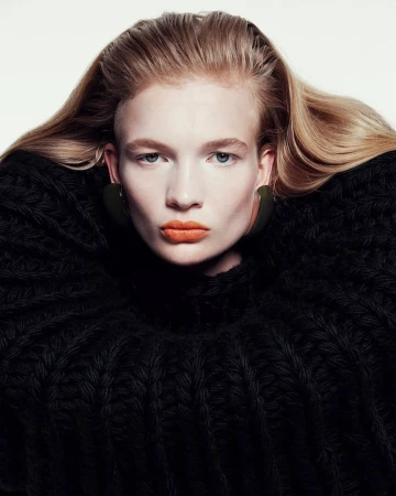 Portrait of a blonde woman wearing a chunky black knit sweater and bright orange lipstick against a white background.