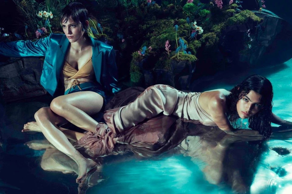 Two fashion models pose in a lush natural setting, one sitting and the other lying in water. The scene features vibrant greens and blues.