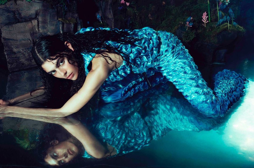 A young woman with dark hair wearing a bright blue ruffled dress laying in shallow water in a dimly lit cave-like environment. Reflection visible.