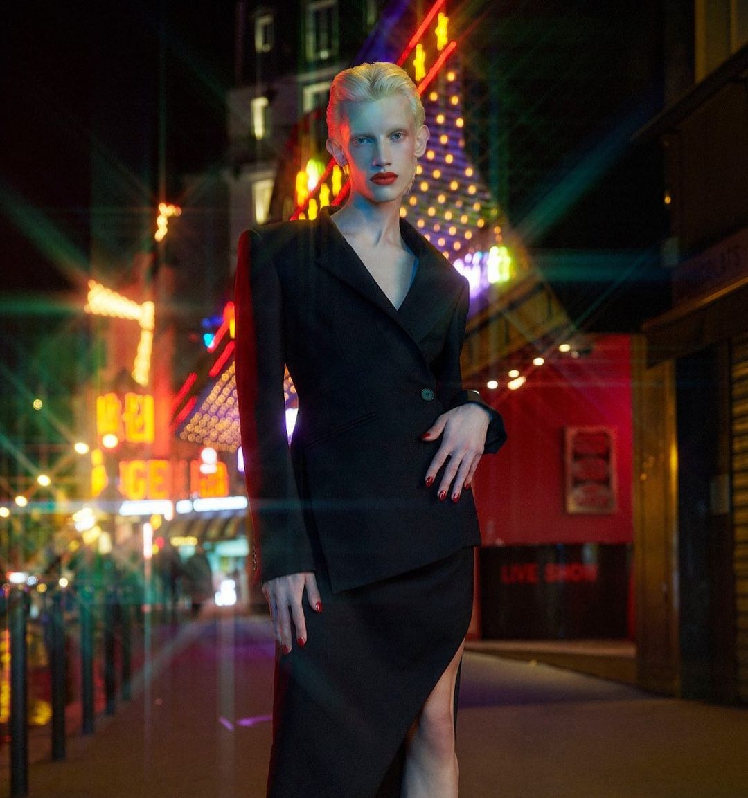 Androgynous model in black suit poses in a neon-lit urban setting at night, featuring a dark street and bright building lights.