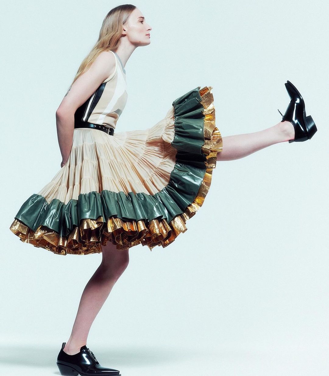 A slender woman with long blonde hair poses in profile, wearing a beige and green tiered ruffled dress and black leather boots against white backdrop.