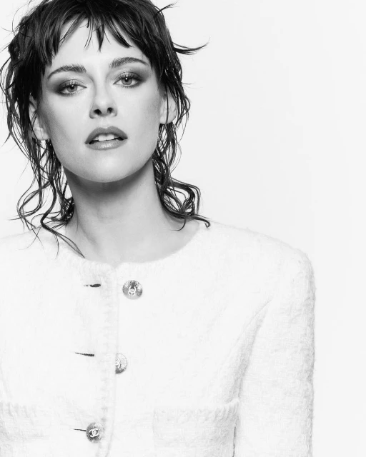 Black and white portrait of Kristen Stewart in a white jacket with short, dark, wet-look hair, posing against a plain background.