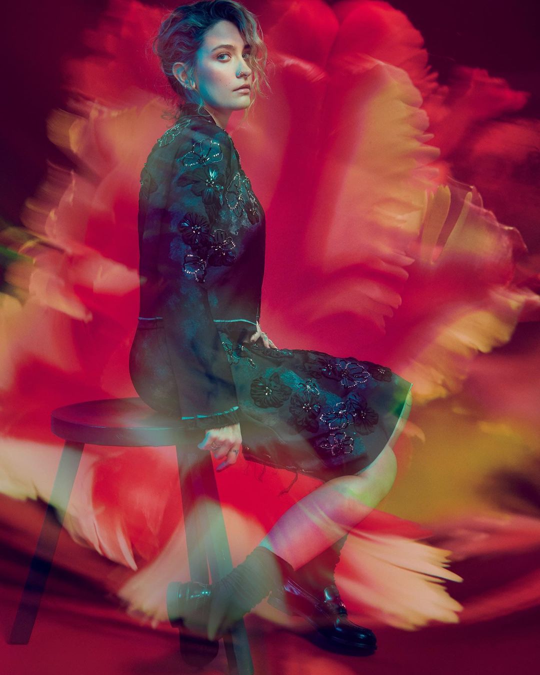 Portrait of adult woman sitting on stool in floral-patterned dress in front of blurred pink floral background in studio lighting.