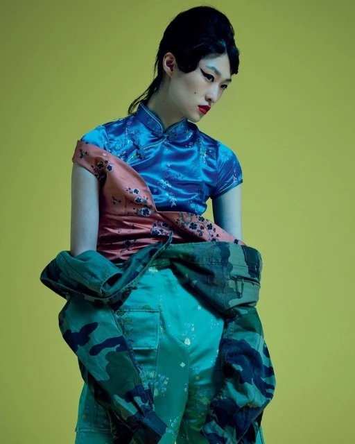 A stylish young woman in a blue, patterned blouse and camouflage pants poses against a yellow backdrop. Fashion editorial shot.