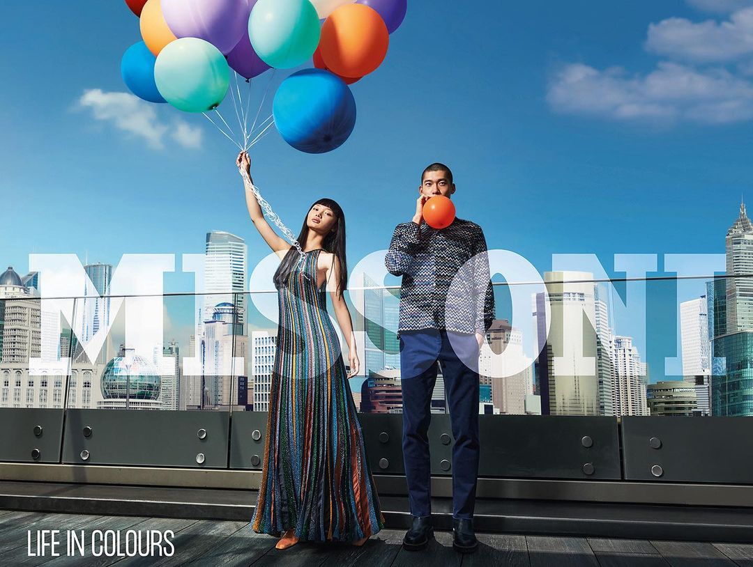 Editorial fashion shot of a man and woman on a rooftop holding balloons against a city skyline, featuring the 'MISSONI - LIFE IN COLOURS' campaign.