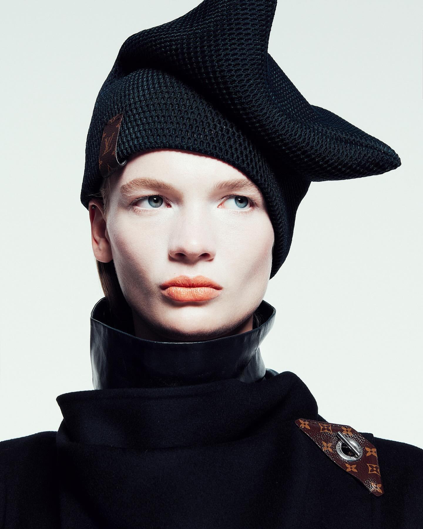 A fashionable, neutral-toned model in a close-up studio portrait wearing a sculptural black knit hat with Louis Vuitton logo detail and a leather-trimmed coat.