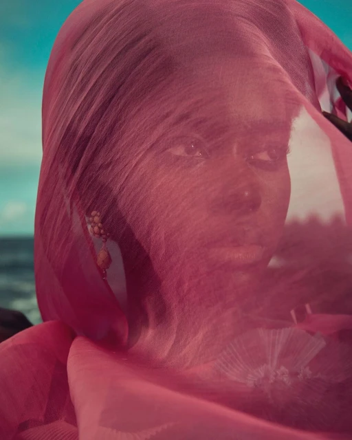 Portrait of a woman with dark skin and a pink veil, set against a backdrop of the ocean and sky on a bright day.