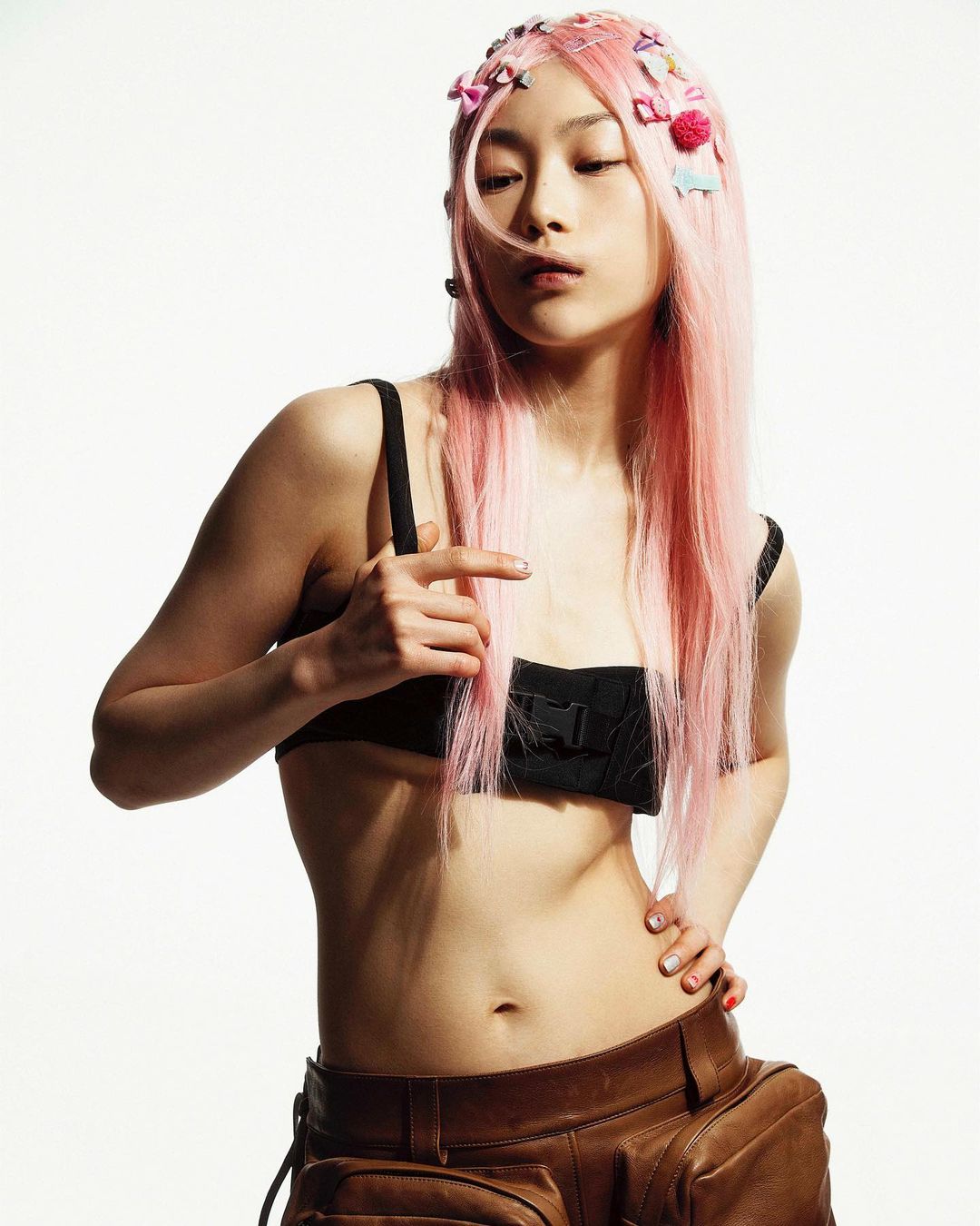 A young Asian woman with pink hair poses in a black bra top and brown leather pants against a bright white backdrop in a studio shot.