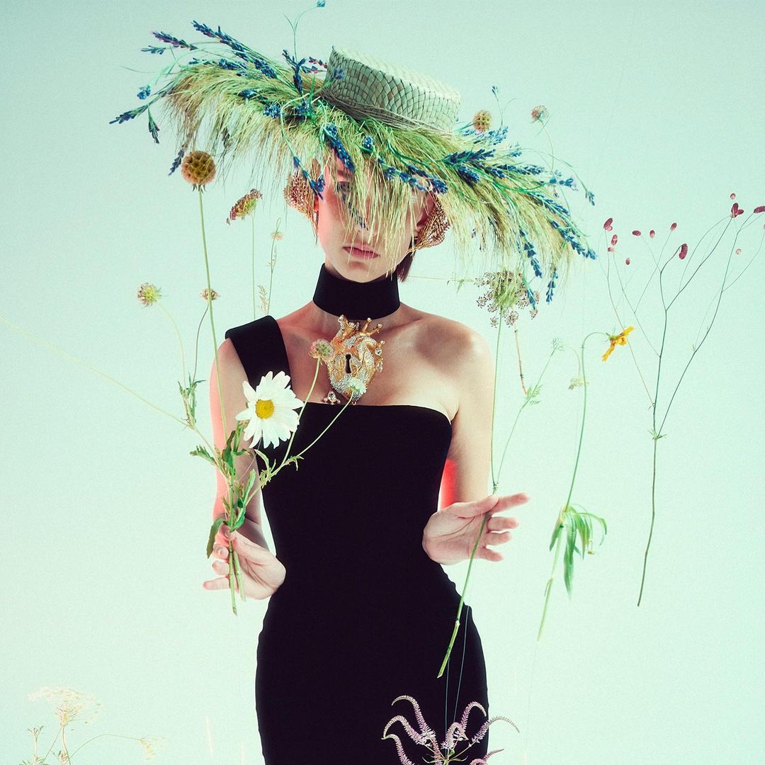 Portrait of a woman in a black dress and a large straw hat adorned with wheat and flowers, set against a neutral-toned background.