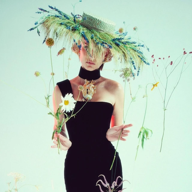 Portrait of a woman in a black dress and a large straw hat adorned with wheat and flowers, set against a neutral-toned background.