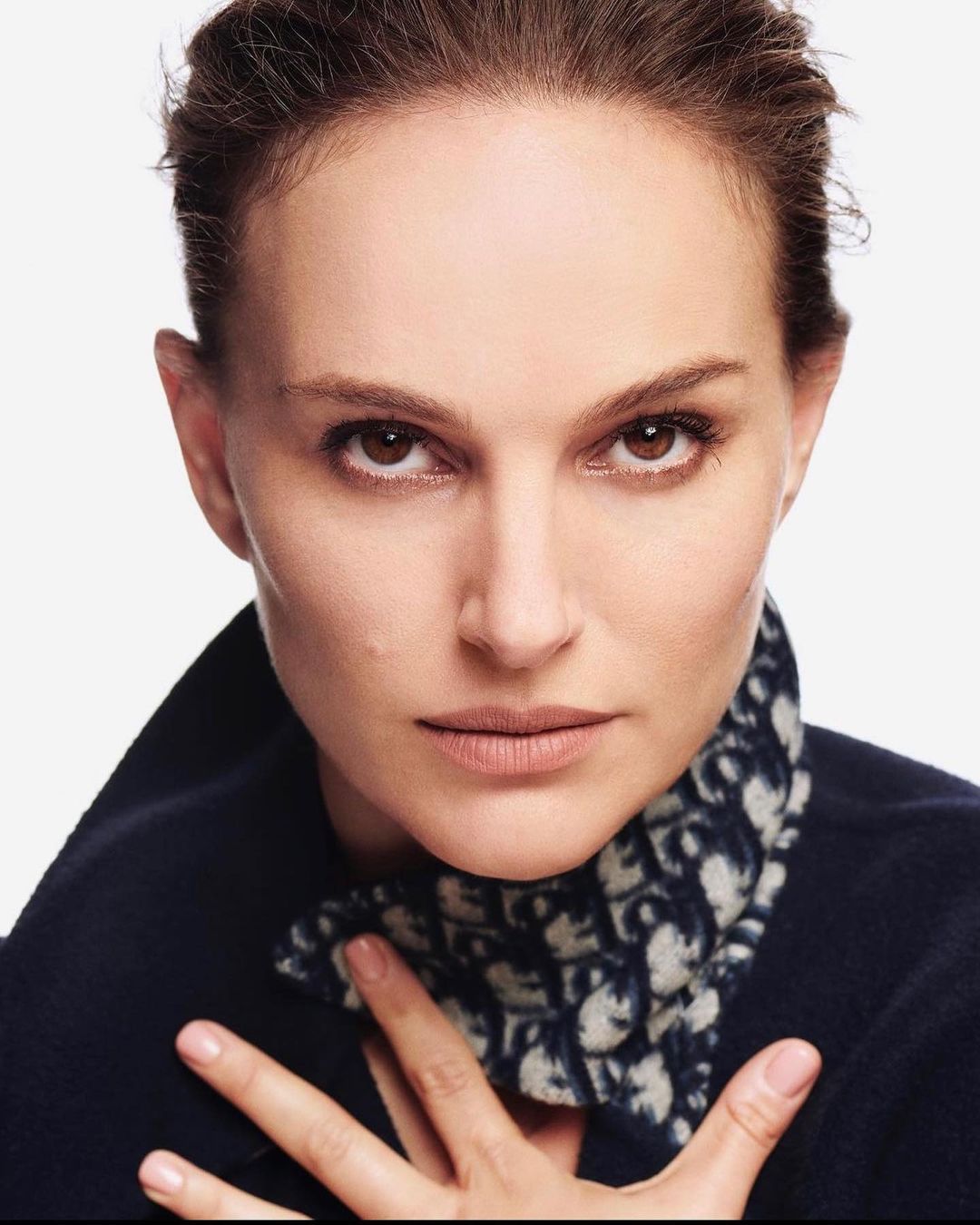 Close-up portrait of a woman with dark hair and brown eyes wearing a dark blue top with patterned collar against a white background.