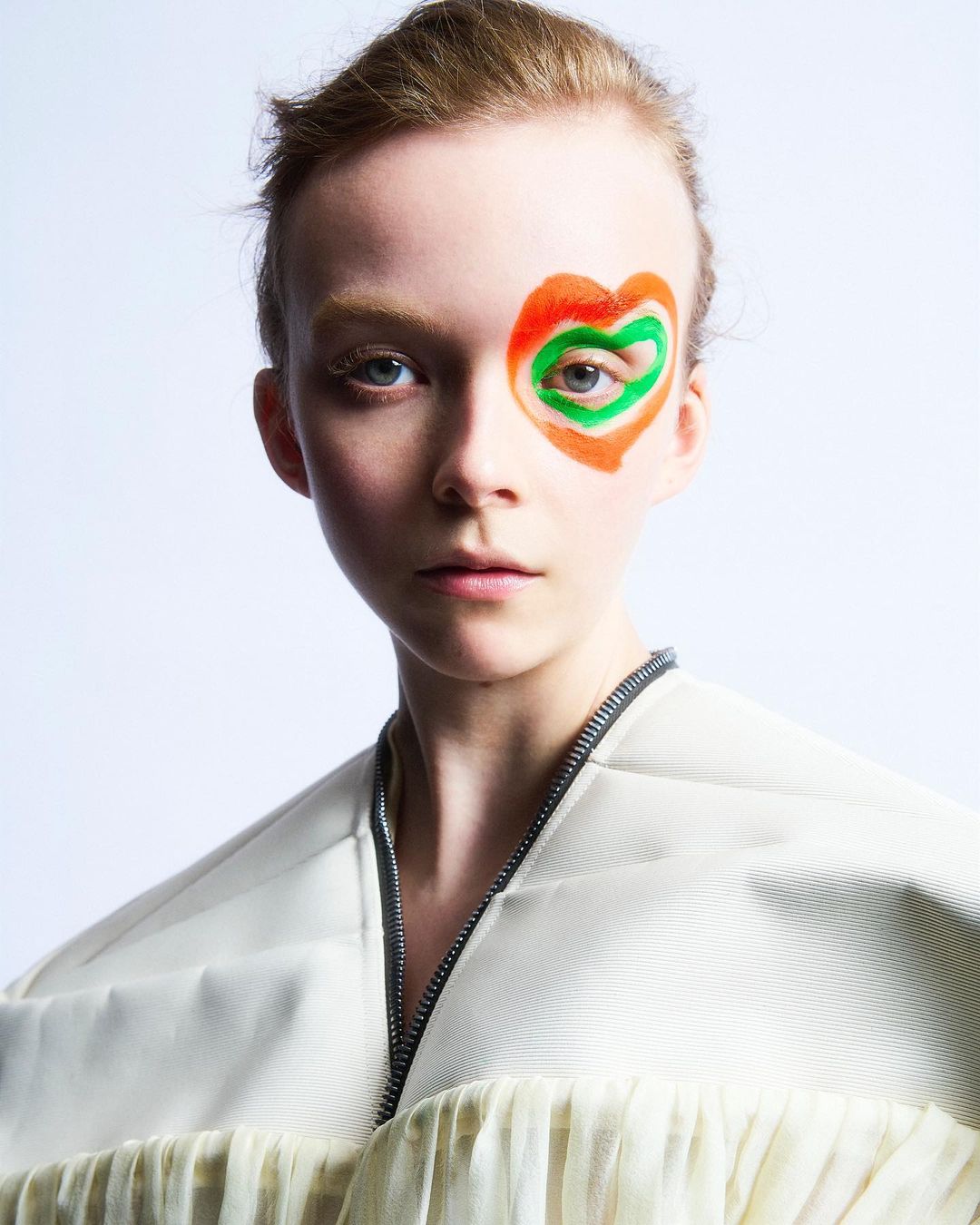 Close-up portrait of a young woman with unique, colorful heart-shaped makeup around her eye, wearing a cream-colored top against white.