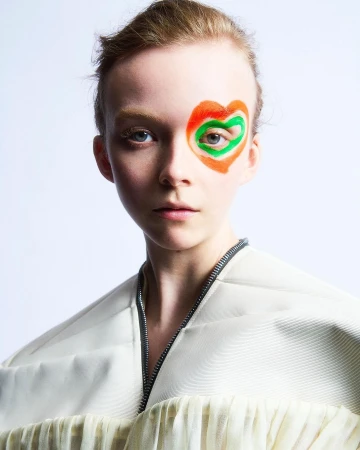 Close-up portrait of a woman with painted heart eye makeup wearing an off-white zipped jacket against a plain background.