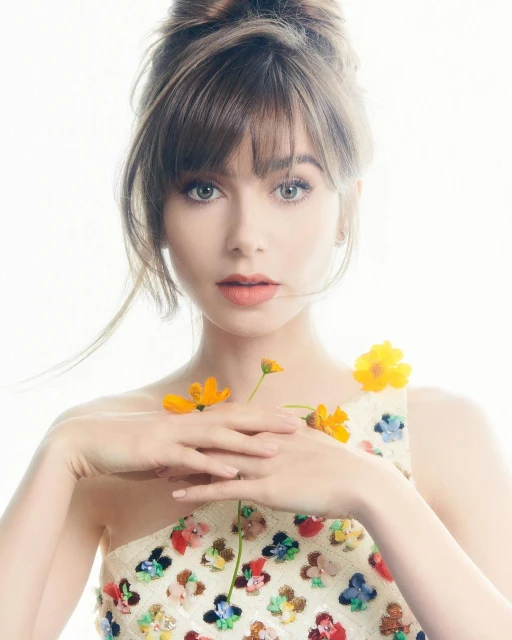 Portrait of a woman with an updo and bangs, wearing a floral-embellished dress and holding a yellow cosmos flower against a white backdrop.