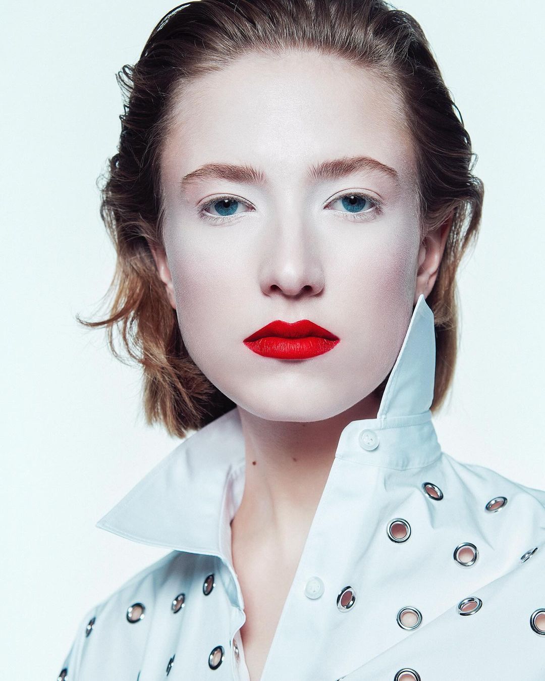 Close-up fashion shot of a fair-skinned woman with bright red lipstick, wearing a white collared shirt with silver grommets, against a white background.