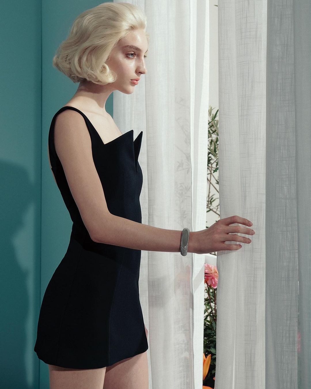 Elegant woman in a short black dress standing near a window with white curtains, looking away, in a fashion editorial setting.
