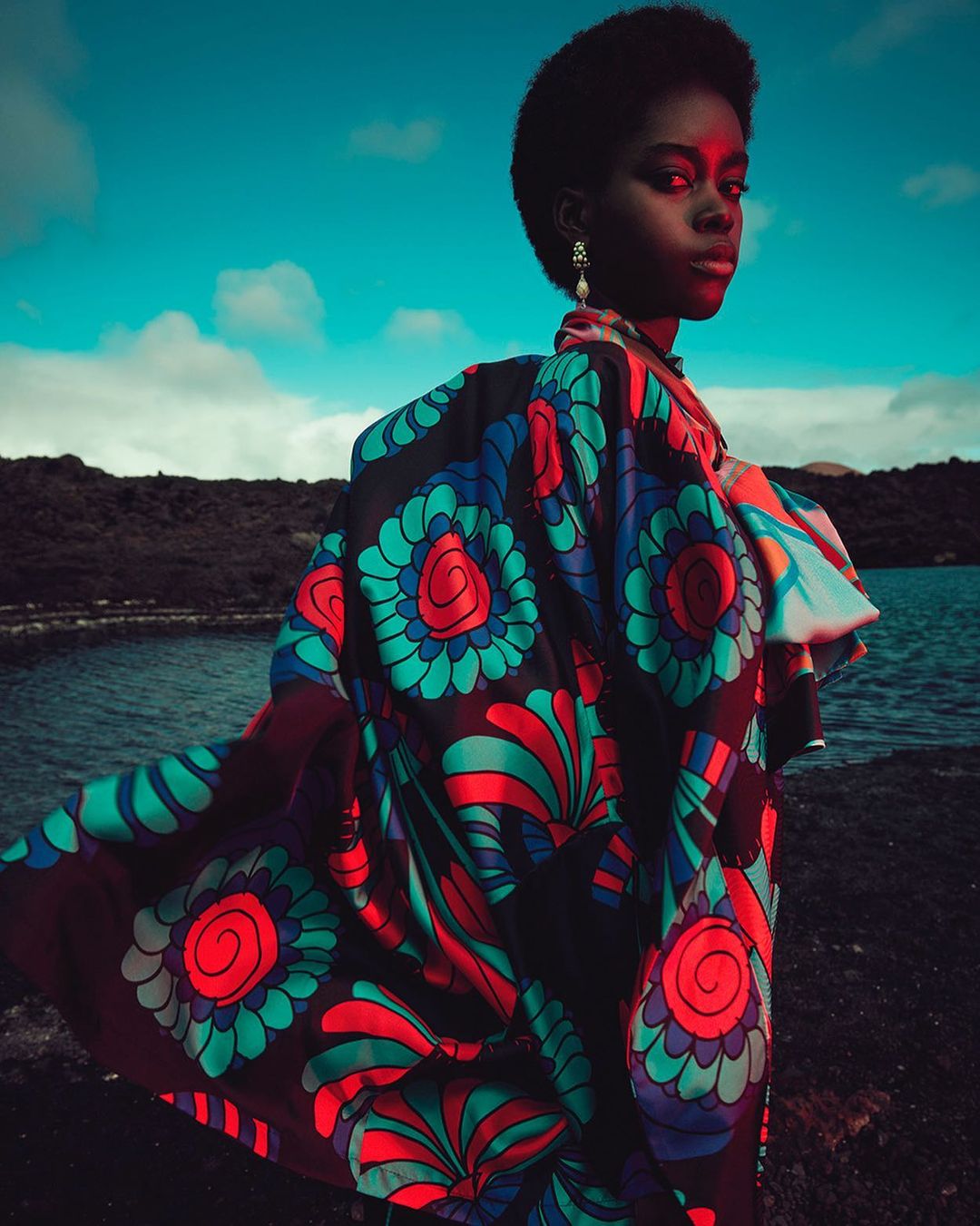 A dark-skinned woman in vibrant floral coat poses outdoors with turquoise sky, body of water, and dark land in the background.