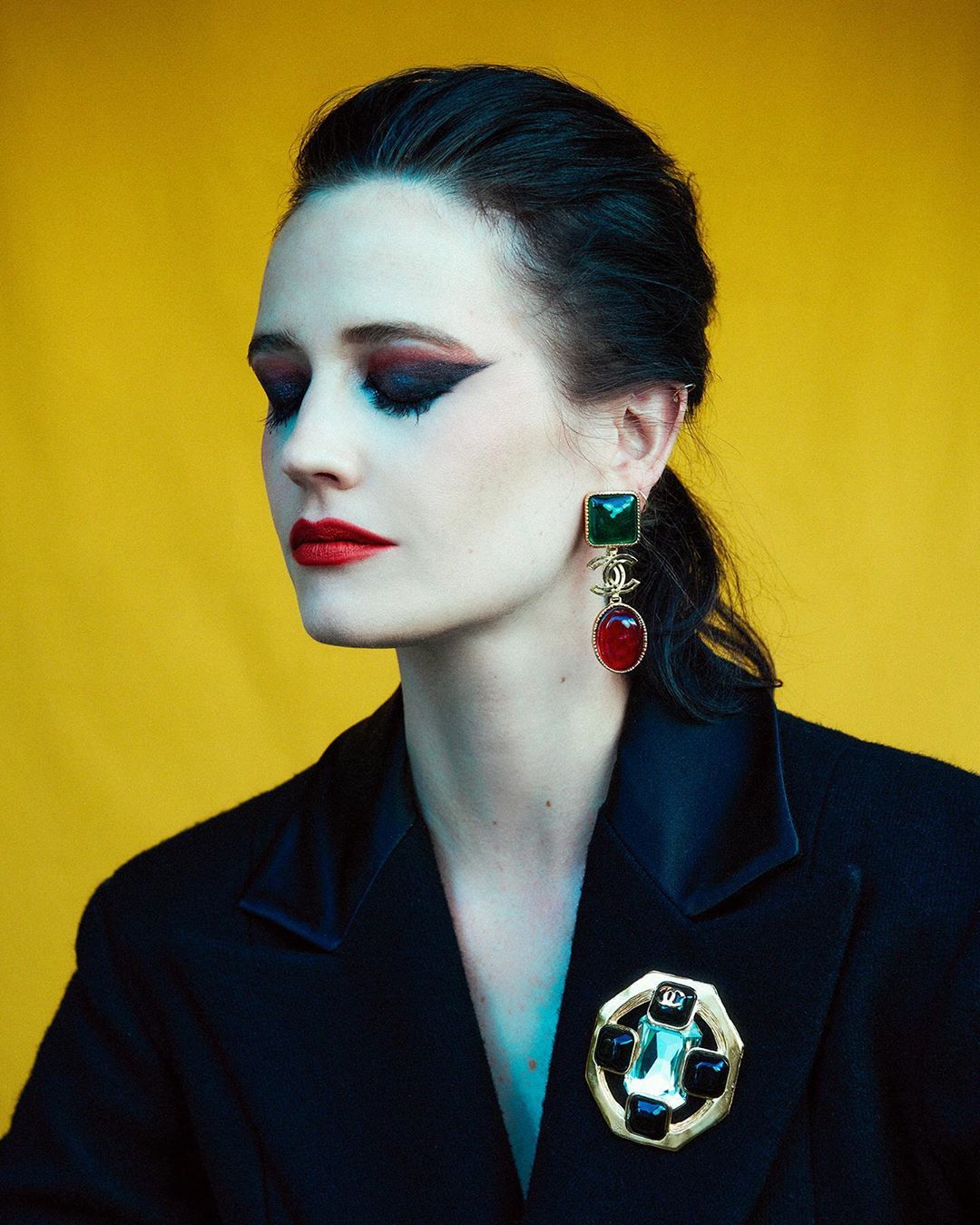 Elegant portrait of a dark-haired woman in black suit, with red lips and heavy eye makeup, wearing gem earrings and brooch against yellow backdrop.