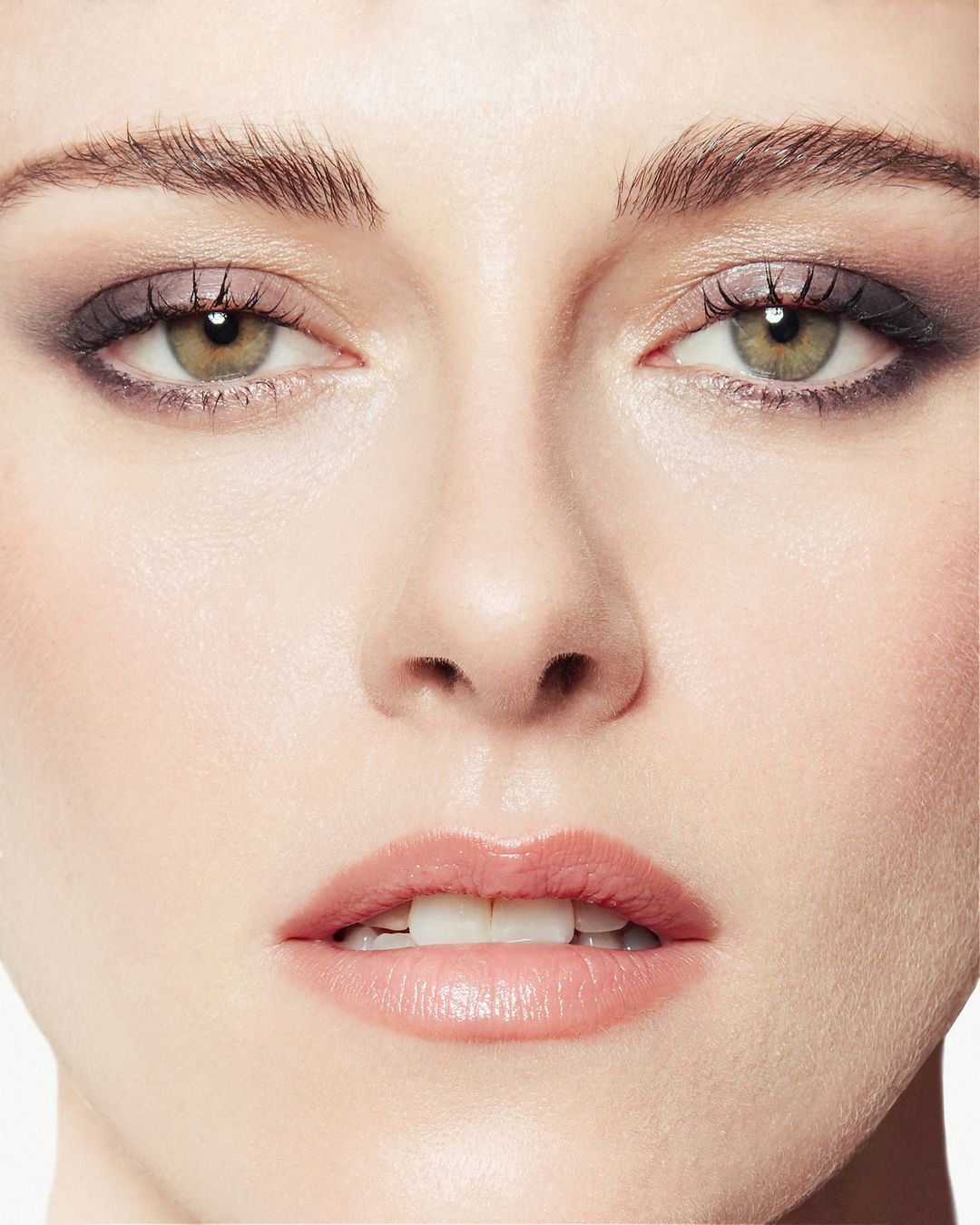 Close-up portrait of a young woman's face with smokey eye makeup, pink lips, and fair skin against a clean, white background in a beauty shot.
