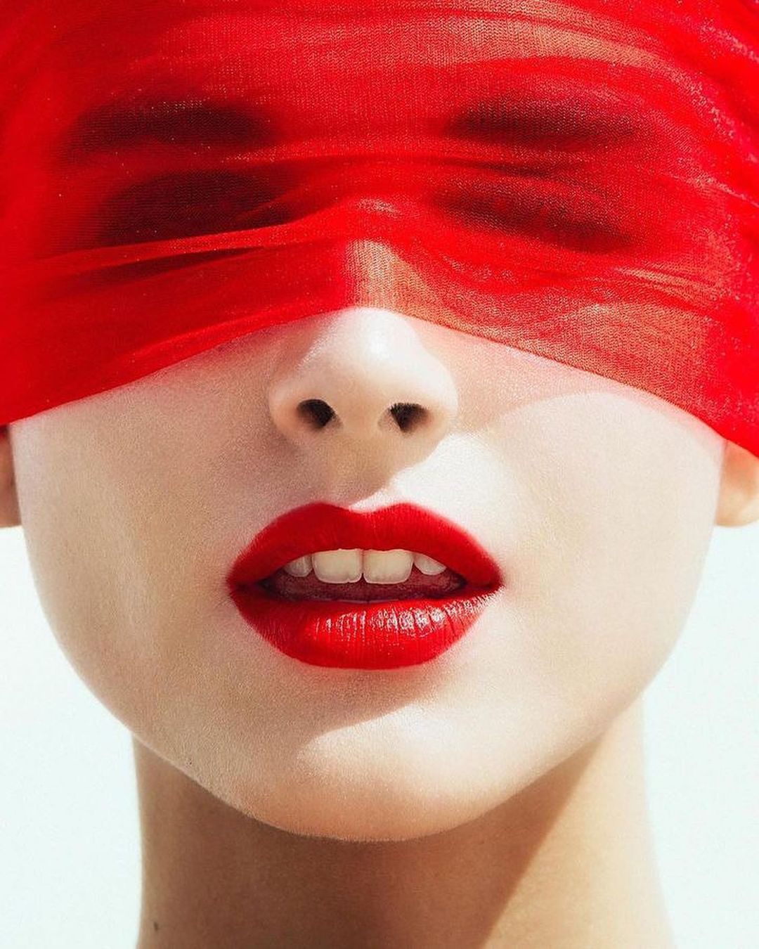 Close-up studio shot of a person with red lips, covered by a red veil. Emphasizes beauty and fashion in a controlled lighting environment.