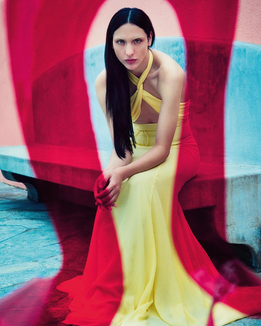 Fashion editorial shot of a slender adult woman with long dark hair, wearing a yellow gown with red color overlay, sitting on a bench.