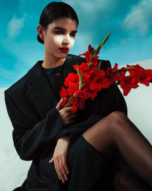 Portrait of a woman in a black jacket holding red gladiolus flowers, set against a bright blue sky with wispy clouds.