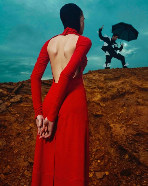 Woman in red dress stands on brown rock formation against a blue sky, with a man in a paper airplane suit holding an umbrella.