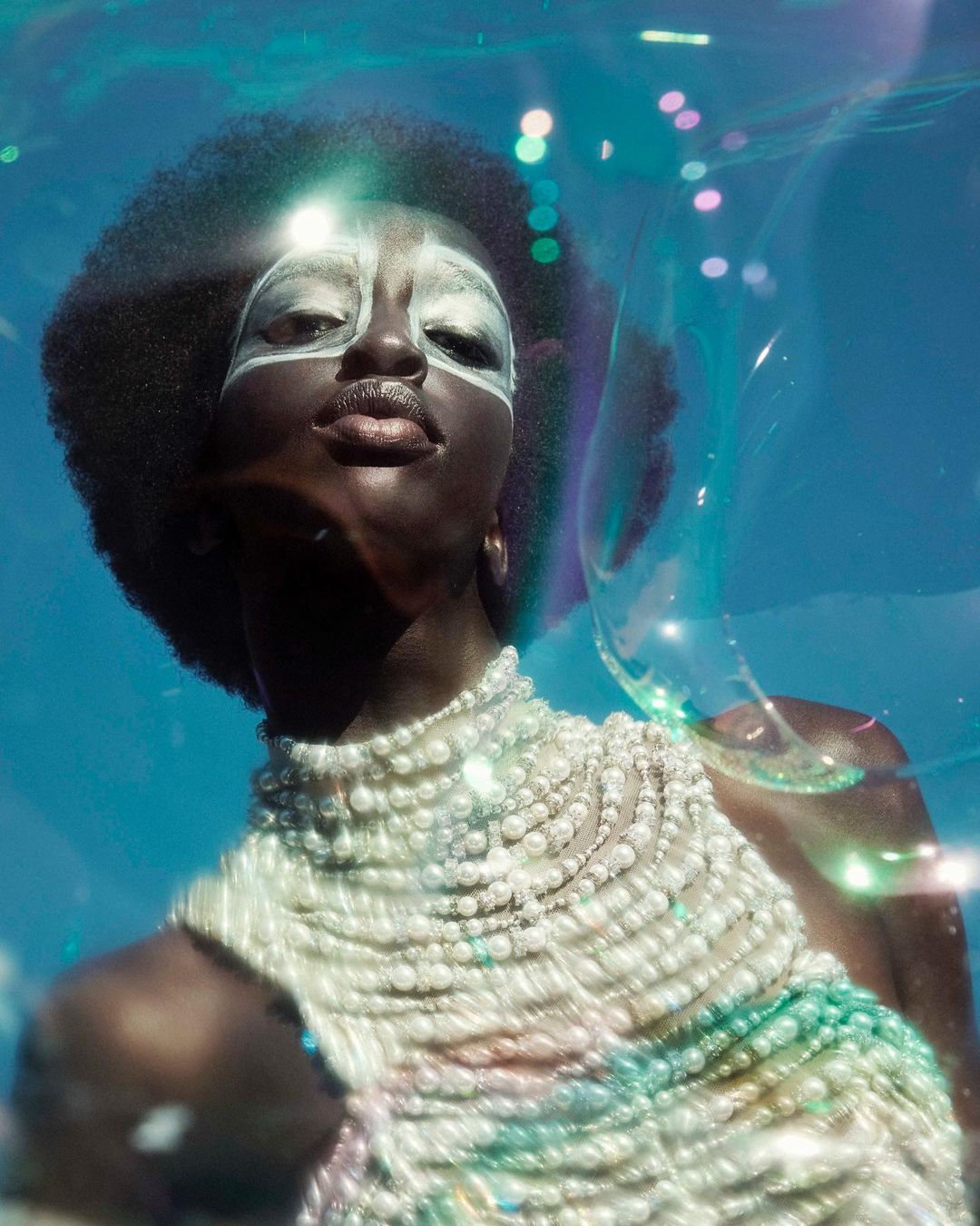 Glamorous portrait of a Black woman with Afro, silver face makeup, and pearl-covered top, posing under water with bubble effects and blue backdrop.