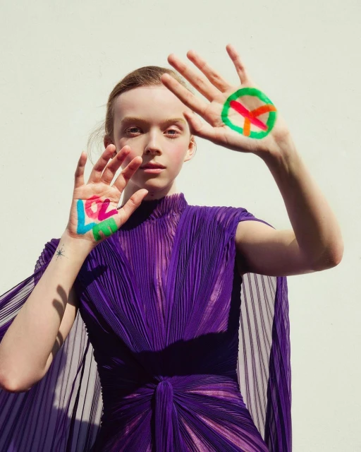 Young adult woman in a purple dress poses with a peace symbol and the word LOVE painted on her hands against a neutral backdrop.