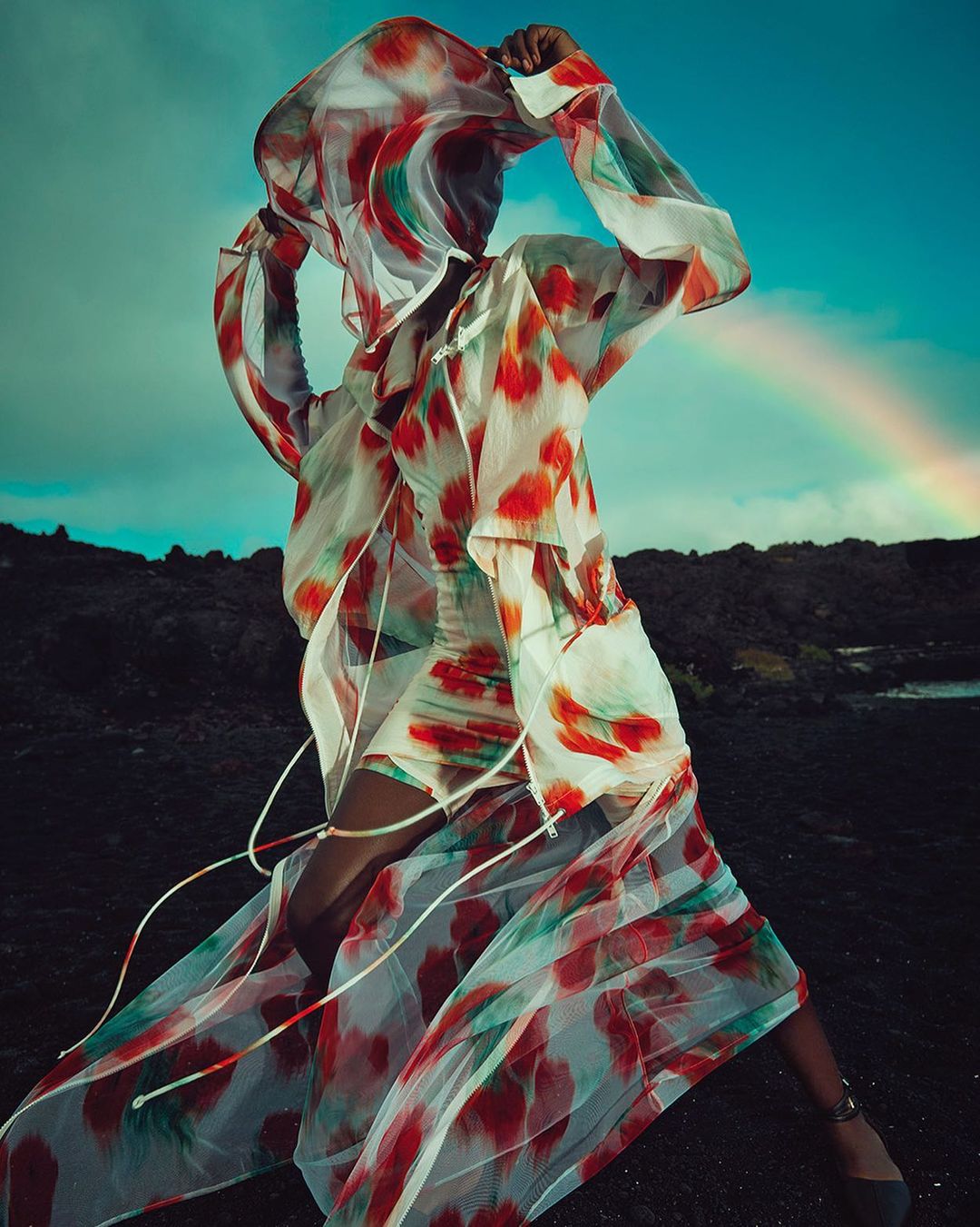 A person wears a white dress with red and green patterns, standing on dark rocky ground under a cloudy sky with a rainbow.