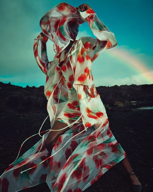 A person wears a white dress with red and green patterns, standing on dark rocky ground under a cloudy sky with a rainbow.