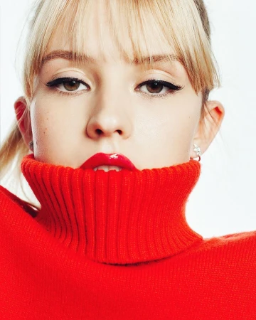 Close-up portrait of a woman with blonde bangs wearing a bright red turtleneck sweater and glossy red lipstick.