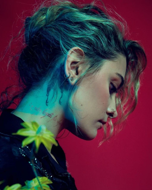 Profile shot of a young woman with flowers, teal and blond hair, wearing a black jacket, against a red studio background.