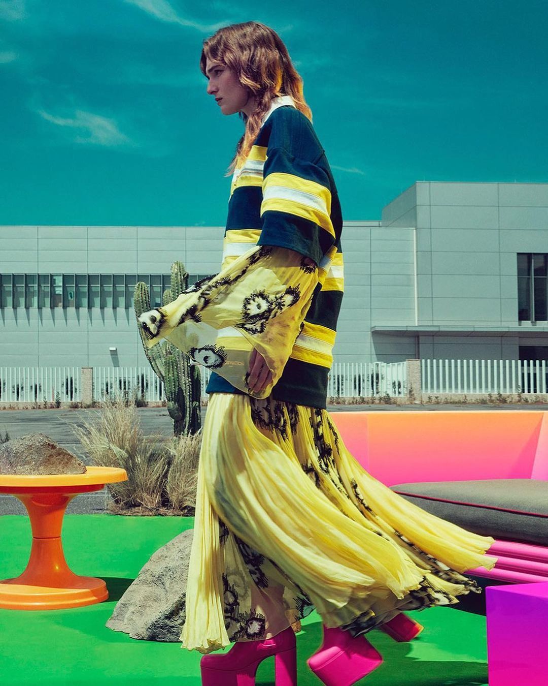 Model in a fashion editorial shoot wearing a striped top, pleated yellow skirt, and pink platform boots against a building backdrop.