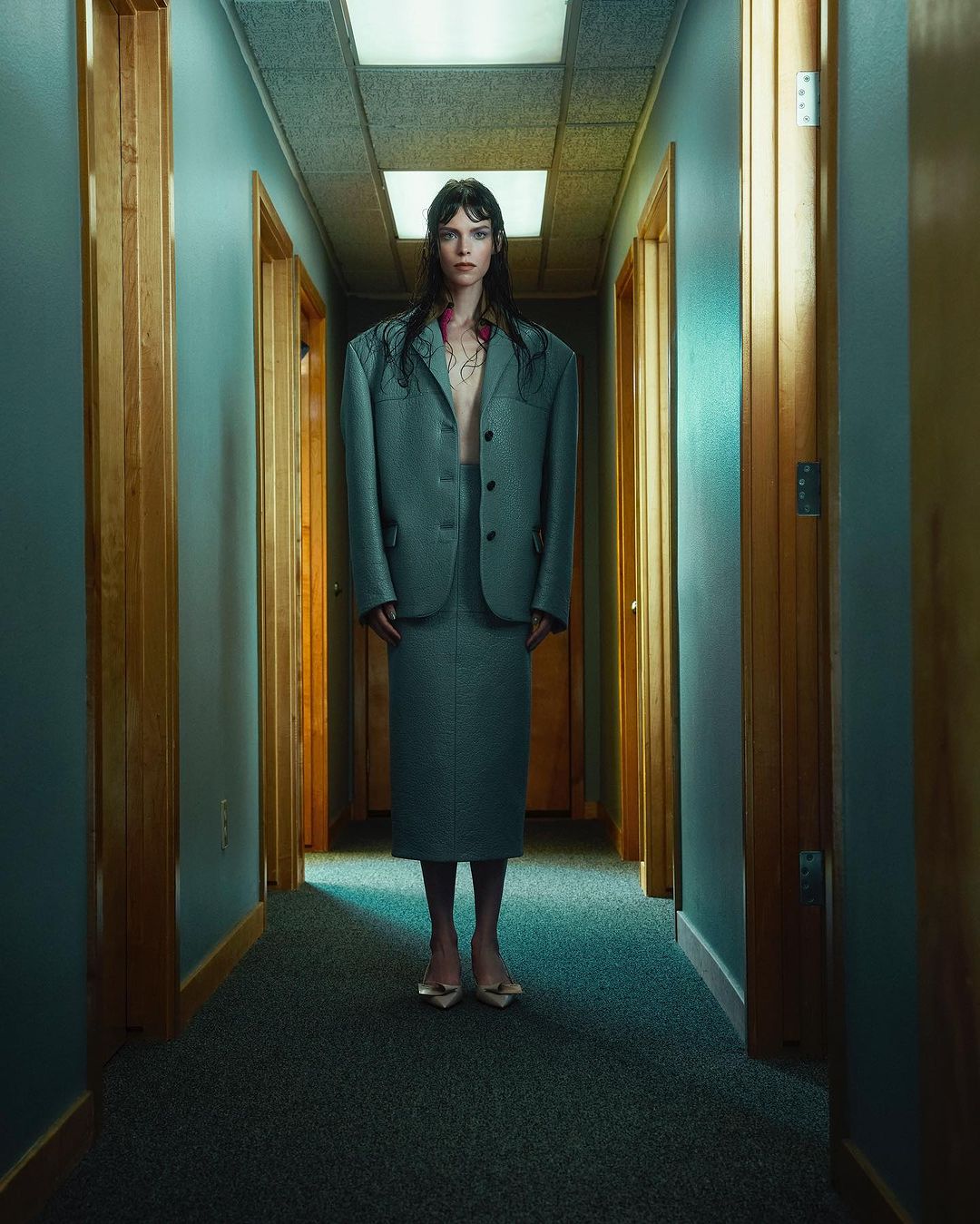 Stylish woman in oversized grey skirt suit stands in a hallway with wood doors and blue walls, creating a surreal fashion portrait.