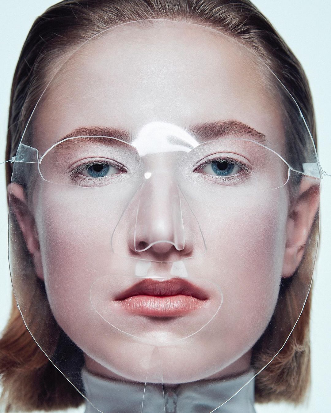 Close-up portrait of a young woman with light skin wearing a transparent face shield, shot in a photo studio against a white backdrop.