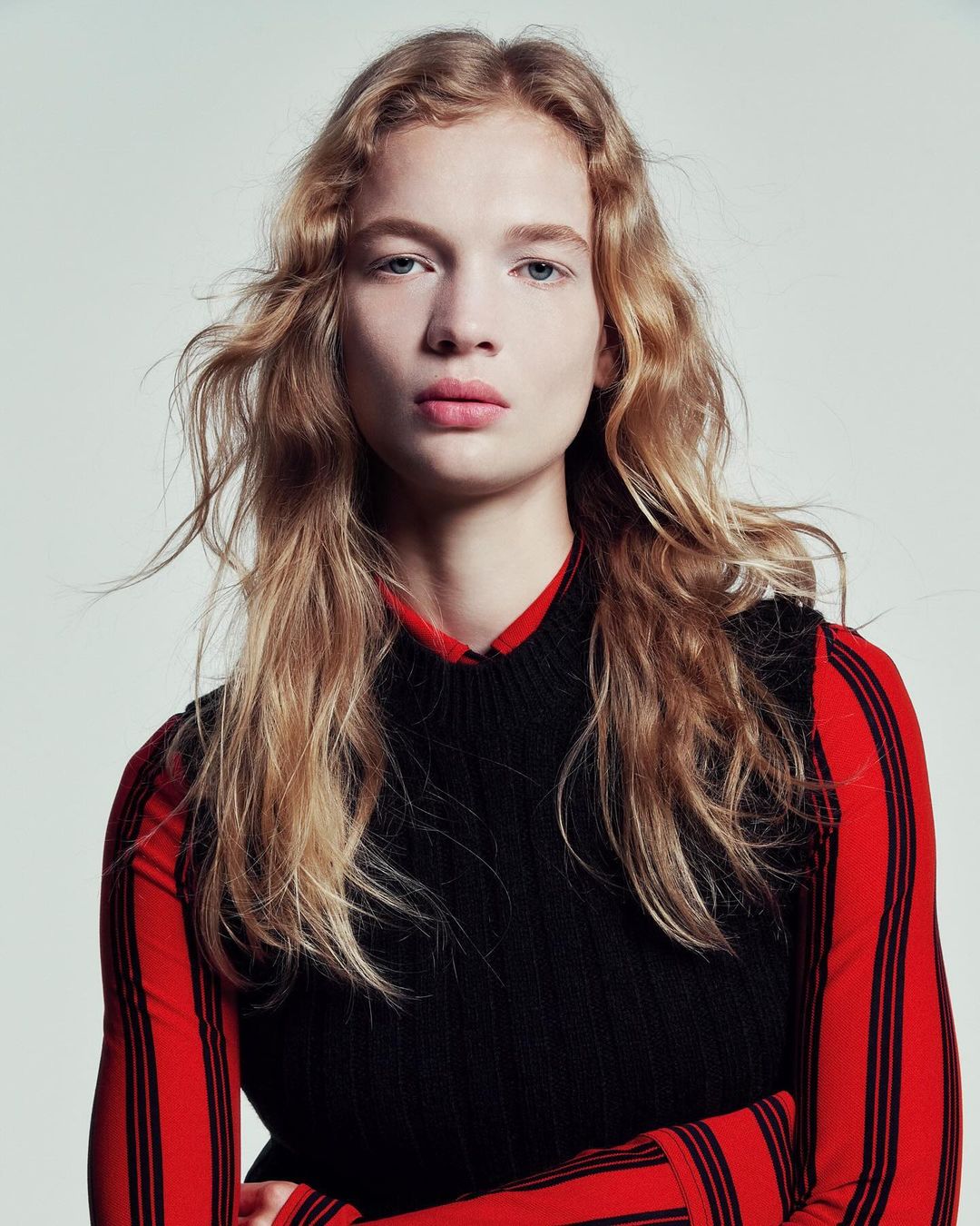 Close-up portrait of a young adult female with long blonde wavy hair, wearing a black sweater vest and a red and black striped shirt.