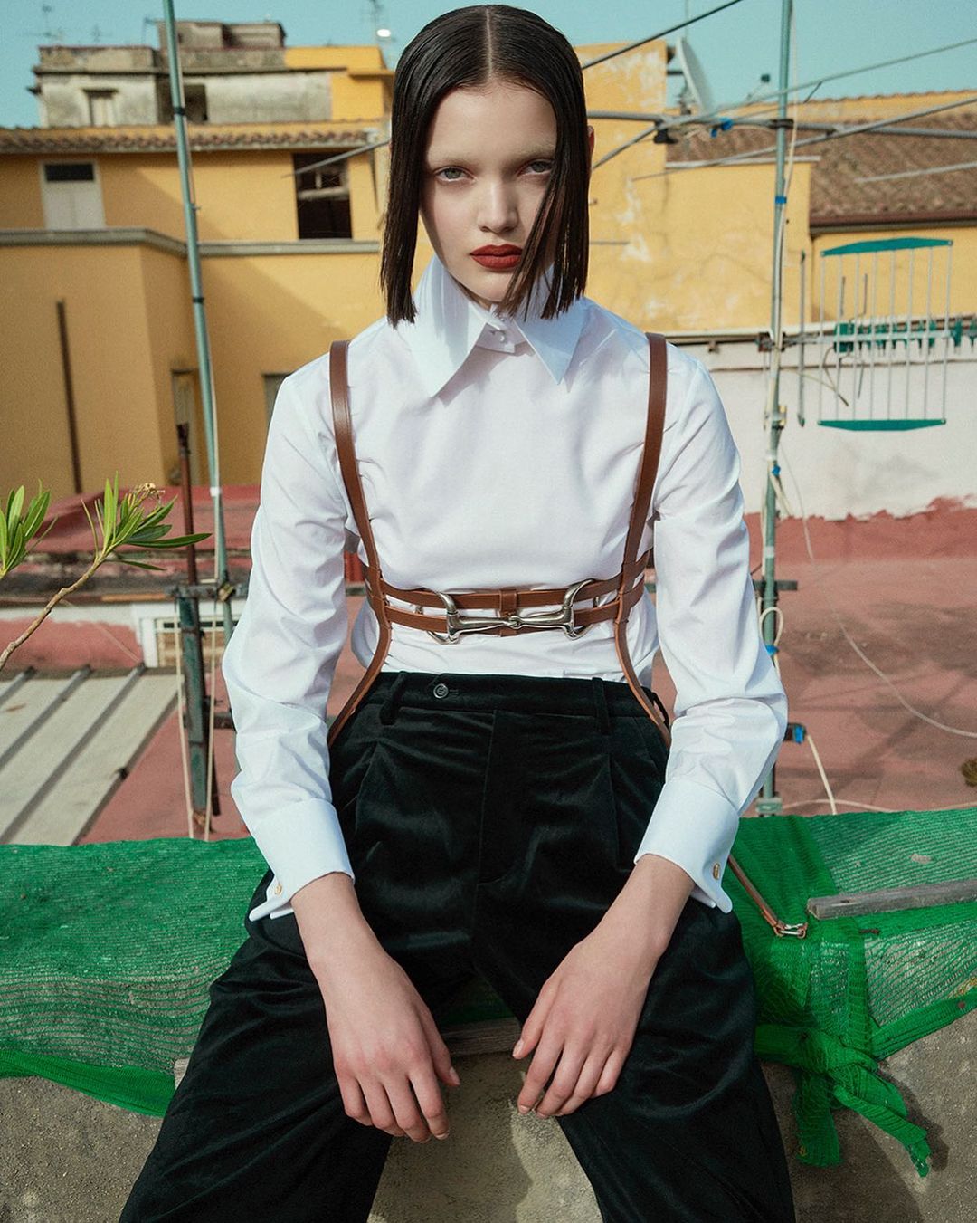 A young female model sits on a roof wearing a white shirt, black trousers, and brown harness. Yellow buildings are in the background.