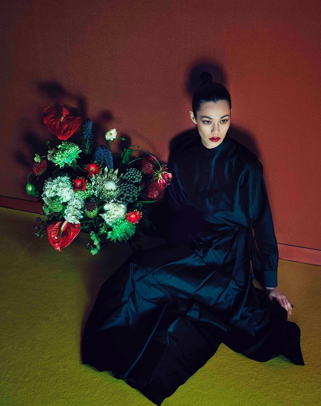 Elegant woman in a dark dress poses with a striking floral arrangement against a warm-toned backdrop, creating a dramatic portrait.