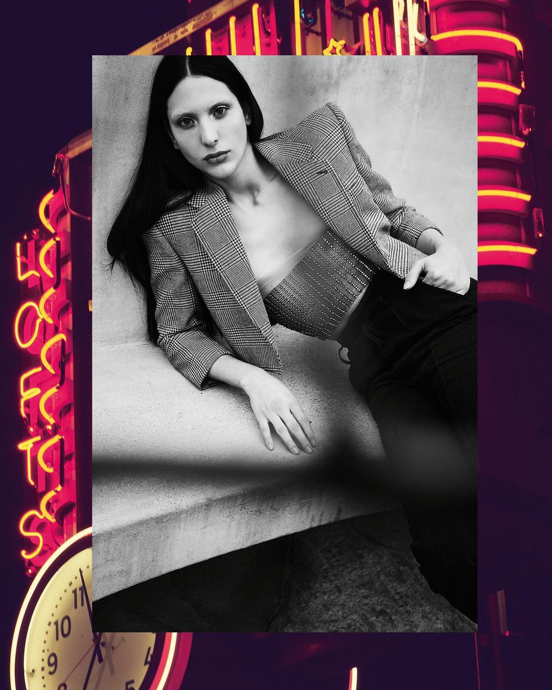 Fashion photo featuring a female model in a blazer and bustier posing in a stylish indoor setting with a neon sign and clock.