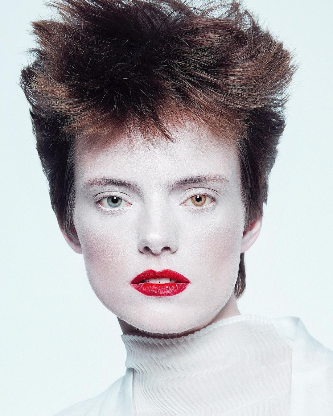 Close-up portrait of a model with white makeup, striking red lips, unique heterochromia eyes, and an edgy, short dark hairstyle.