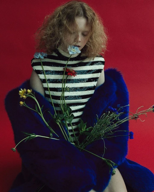 A young person with curly hair poses indoors in a striped top and blue coat, holding flowers against a red backdrop in a studio shot.