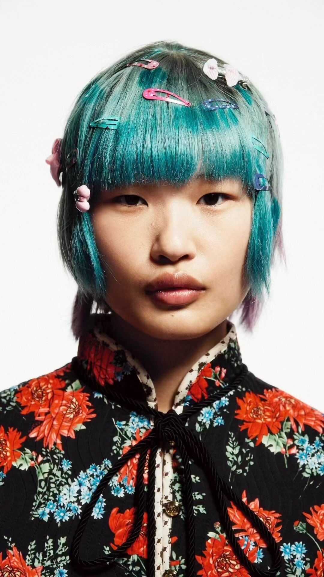 Close-up portrait of a young Asian woman with blue hair adorned with colorful hair clips. She wears a floral top against a white background.