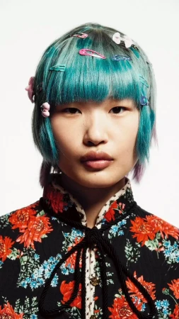 Close-up portrait of a young Asian woman with blue hair adorned with colorful hair clips. She wears a floral top against a white background.