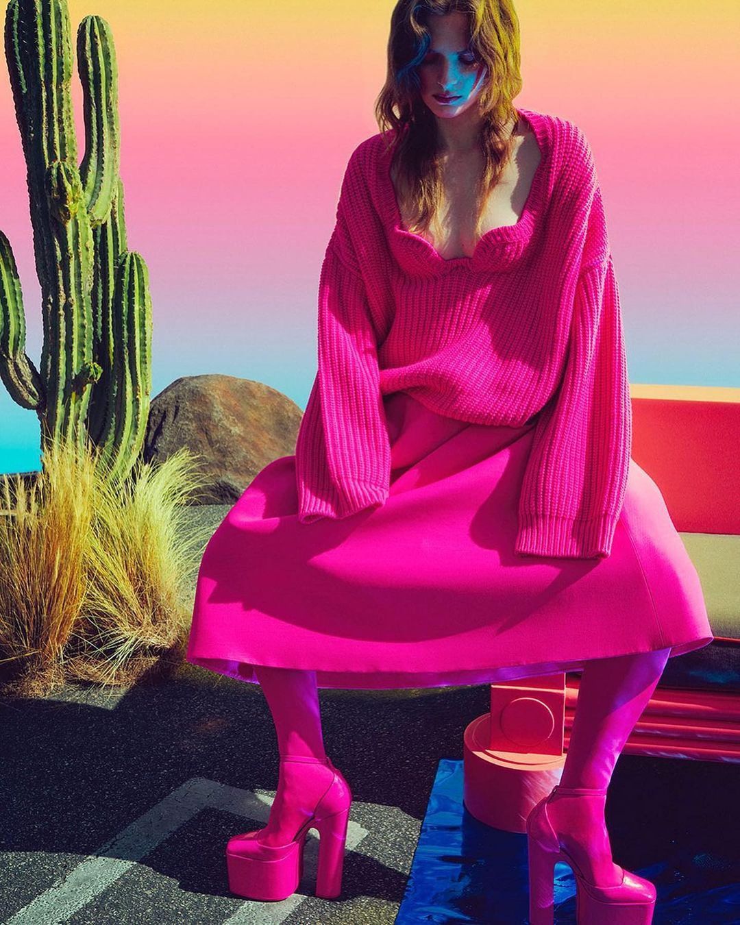 Woman in pink sweater and platform heels poses by cactus in vibrant, colorful landscape. Fashion-forward, expressive, and visually striking.
