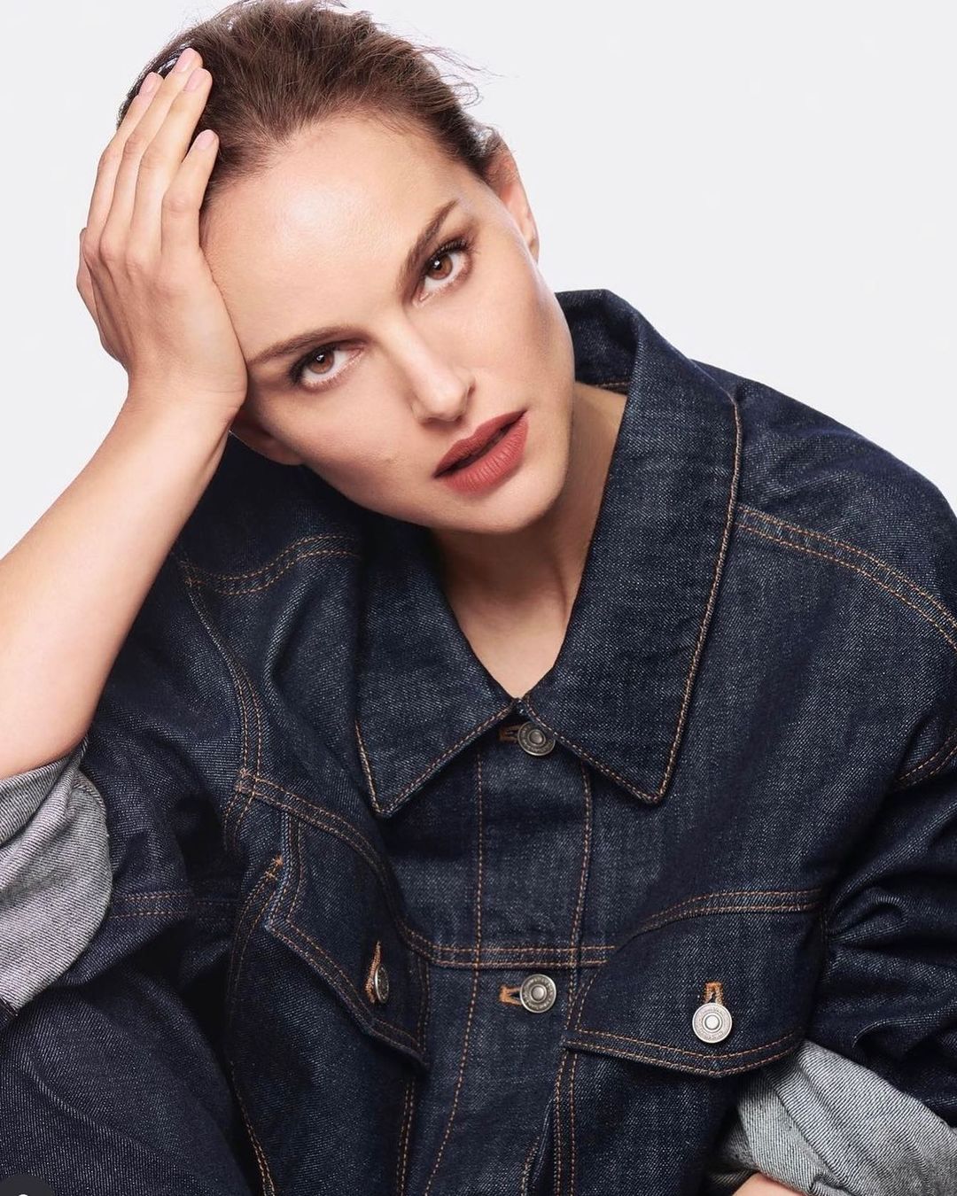 Portrait of a woman with dark hair in a denim jacket against a white background, looking directly at the camera with a neutral expression.