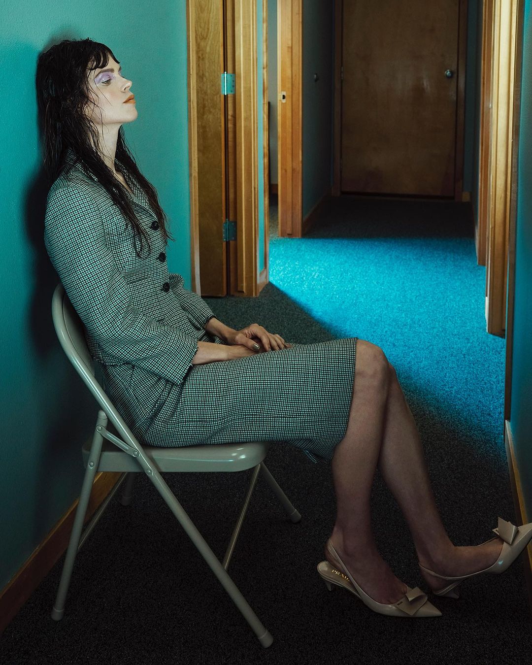 A young woman poses in a houndstooth skirt suit on a folding chair in a hallway with wood doors under bright, directional lighting.