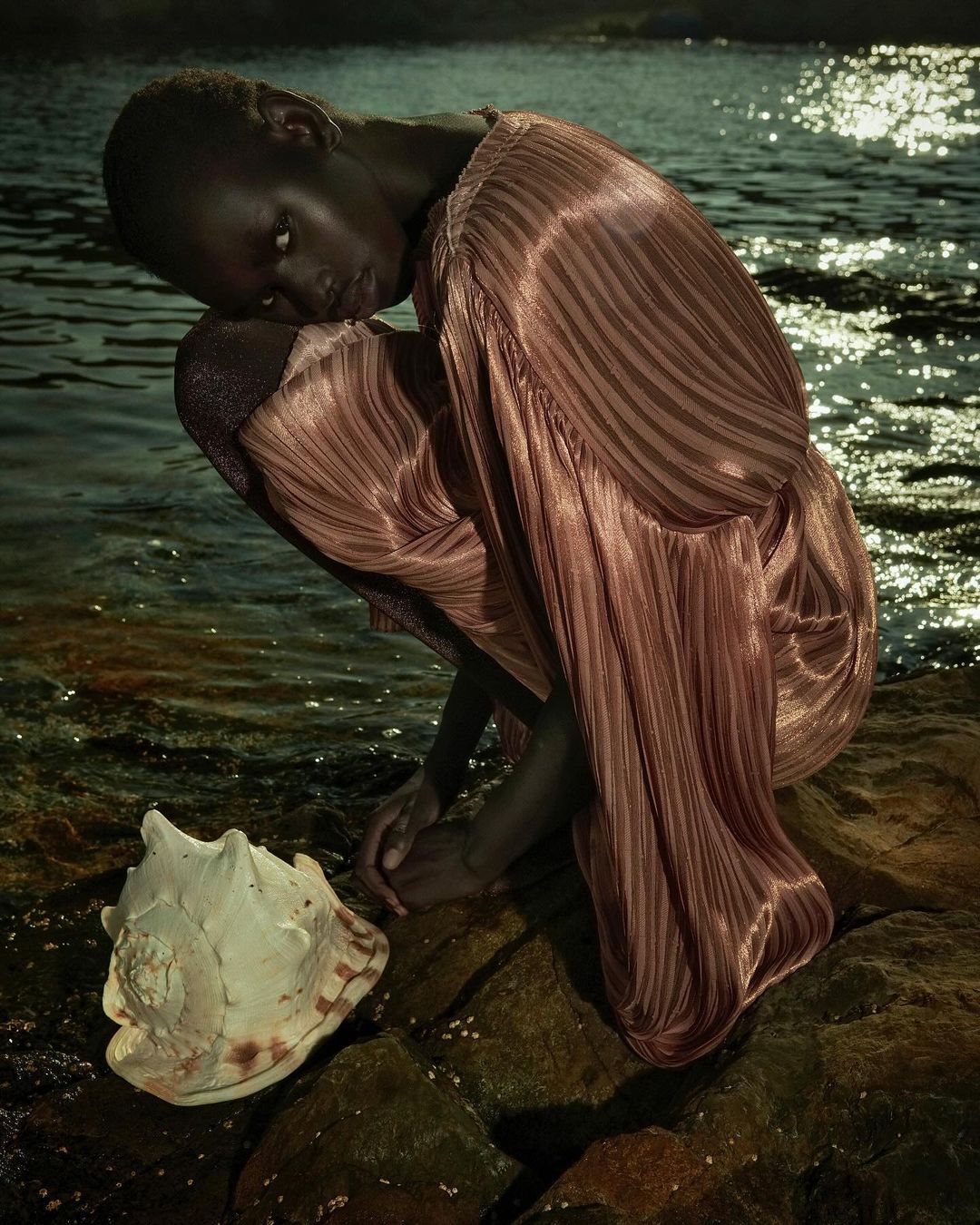 Woman in pleated rose gold dress poses crouching on rocks by water with seashell in a nighttime outdoor fashion photography.