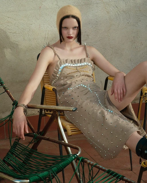 A model sits on a yellow chair outdoors wearing a tan dress and hat, showcasing fashion and style in a relaxed pose against a plain wall.