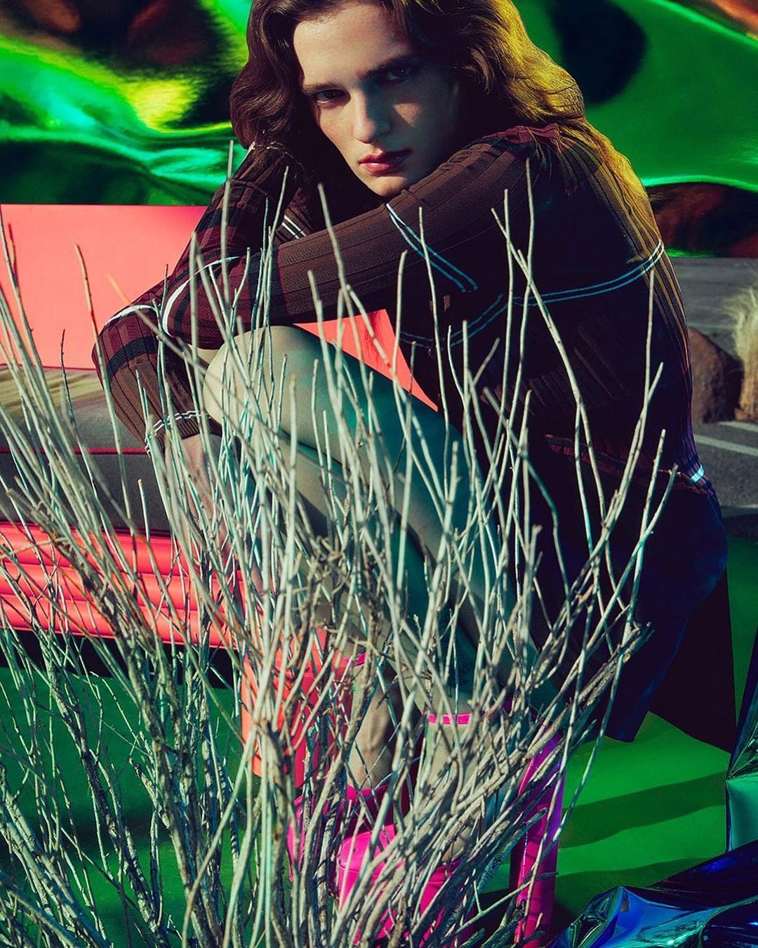 Model in stylish sweater and pink boots poses crouching behind white branches against a vibrant backdrop of green and coral hues.